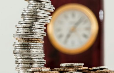 A close-up image of stacked coins with a blurred clock, symbolizing time and money relationship.